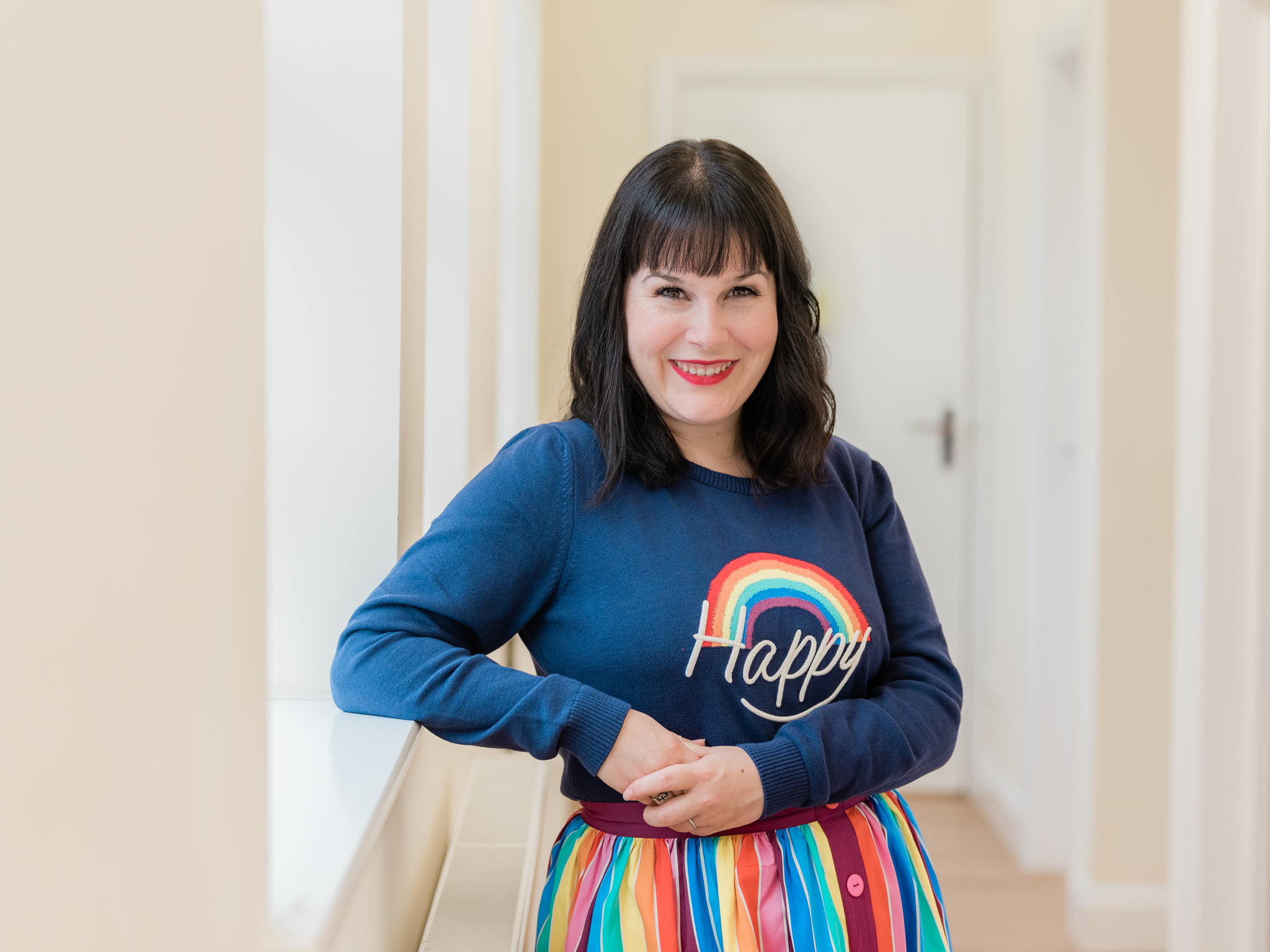Life Coach Miranda Dawe stands smiling, wearing a rainbow top that says, 'happy'.